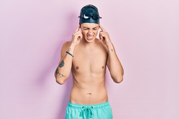 Young hispanic man wearing swimwear and swimmer glasses covering ears with fingers with annoyed...