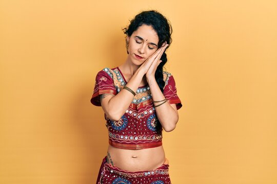 Young woman wearing bindi and bollywood clothing sleeping tired dreaming and posing with hands together while smiling with closed eyes.