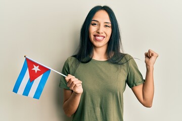 Young hispanic girl holding cuba flag screaming proud, celebrating victory and success very excited...
