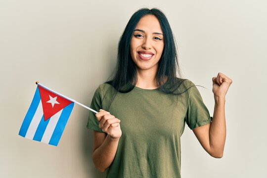Young hispanic girl holding cuba flag screaming proud, celebrating victory and success very excited with raised arm
