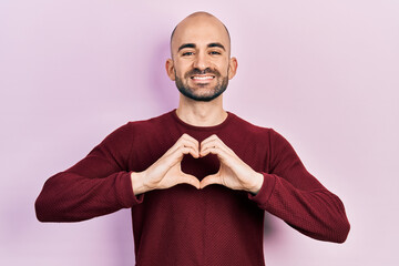 Young bald man doing heart symbol with hands smiling with a happy and cool smile on face. showing teeth.