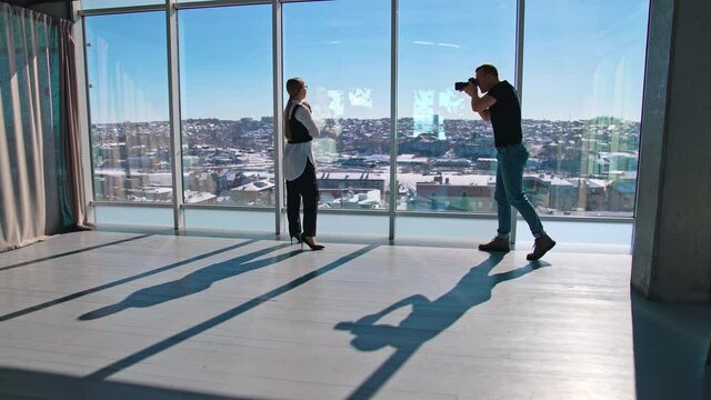 Male photographer taking photos of a young woman. Business woman posing on camera in the light office room. Panoramic windows view.