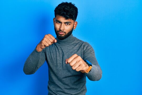 Arab man with beard wearing turtleneck sweater punching fist to fight, aggressive and angry attack, threat and violence