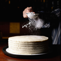 Crop baker sprinkling sugar powder through strainer on whole carrot cake placed on table in dark...