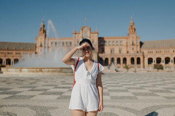 Young tourist woman with glasses wearing a white dress and red backpack is smiling and covering her eyes from the sun while standing in the Plaza de España of Seville. Blinded by the harsh sun.