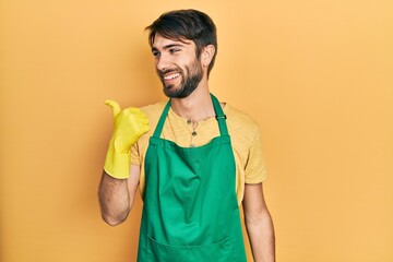 Young hispanic man wearing cleaner apron and gloves pointing thumb up to the side smiling happy with open mouth