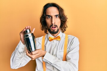 Young hispanic man preparing cocktail mixing drink with shaker in shock face, looking skeptical and sarcastic, surprised with open mouth