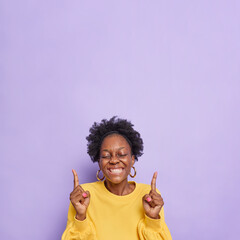 Cheerful overjoyed dark skinned young woman shows cool advertisement rejoices something points at sale banners or promo deals smiles broadly wears yellow jumper isolated over purple background