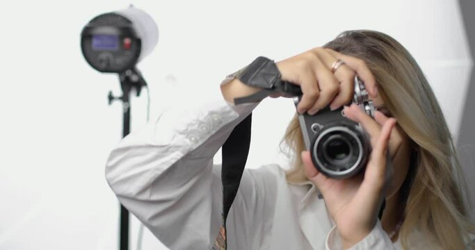 Portrait of a stylish young woman photographer in a white shirt with a pierced nose and long blond hair, making shots. Smiling. Equipment light in the background. Professions, hobbies, the arts.