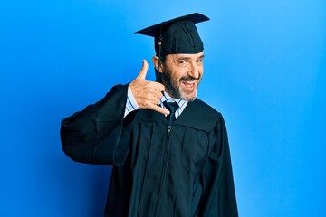 Middle age hispanic man wearing graduation cap and ceremony robe smiling doing phone gesture with hand and fingers like talking on the telephone. communicating concepts.