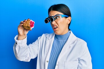 Beautiful brunette jeweller woman holding geode stone wearing magnifier glasses clueless and confused expression. doubt concept.