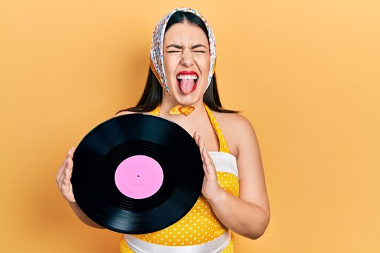 Young hispanic woman wearing pin up style holding vinyl disc sticking tongue out happy with funny expression.
