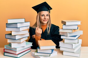 Young caucasian woman wearing graduation ceremony robe sitting on the table beckoning come here gesture with hand inviting welcoming happy and smiling