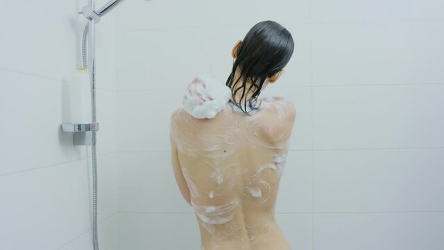 Young caucasian woman with dark hair using shower gel and shampoo while taking shower at home. Back view of naked wet lady. Freshness concept.
