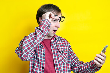 handsome surprised and excited man with open mouth in red checkered shirt taking off glasses on forehead and looking at smartphone on yellow background with copy space