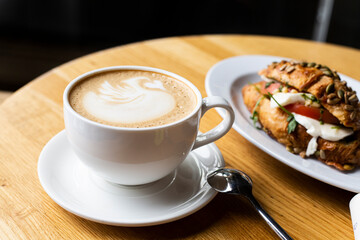 a cup of cappuccino in a cafe stands on a wooden table. in the background croissant with tomatoes