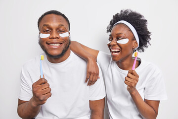 Studio shot of positive Afro American couple have fun undergo daily hygiene routines pose withh toothbrushes take care of teeth apply beauty patches under eyes to remove fine lines and bags.