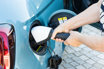 The hand of a Caucasian woman inserts a CCS charging plug into the connector of a blue electric vehicle