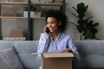 Millennial mixed race woman sit on cozy couch at home prepare cardboard box to send gift to friend call by phone order courier delivery service. Young black female talk on cell unpack parcel container - Powered by Adobe