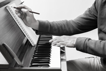 black and white male songwriter writing a song on music sheet while playing chord on acoustic piano...
