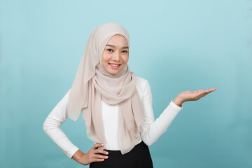 Portrait of smiling Asian young Muslim woman in hijab head scarf standing and raising her hands against blue colour background