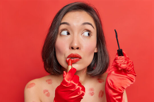 Beauty makeup. Asian lady applies lipstick and mascara uses decorative cosmetics for fabulous look wears red gloves poses undressed against bright studio background gets ready for special occasion