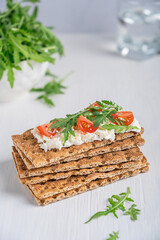Stack of crispbread - a flat and dry type of crispy cracker, made of whole wheat flour with ricotta, cherry tomato slices and rocket leaf topping on white wooden background at kitchen. Vertical