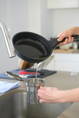 Woman pouring used cooking oil from an old frying pan into a metal cooking container in her kitchen.