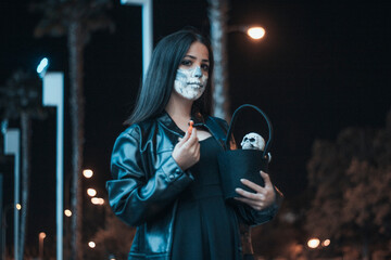 Fototapeta na wymiar Chica joven de pelo castaño vestida de chaqueta disfrazada en la oscuridad para celebrar el dia de Halloween con su cesta de caramelos