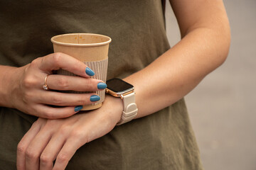 girl holds a disposable paper cup with coffee next to a smart watch on her hand, break time on the street in a summer park, drink coffee on the street