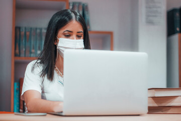 Chica joven con mascarilla estudiando para el examen en la biblioteca del campus universitario con libros y ordenador portatil laptop