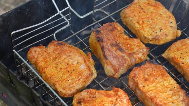 Close-up view 4k stock video footage of cooking tasty juicy pork meat steaks laying on grill grid on open fire over flaming, burning and steaming with smoke black organic wooden charcoal in pit