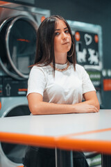 Adolescente deprimida con pendientes y pelo castaño esperando junto a la mesa de la lavanderia en una tienda de lavado de lavadoras para lavar su ropa 