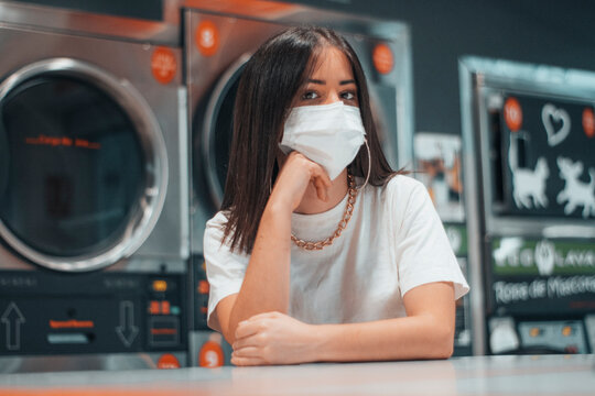 Se&ntilde;ora joven de pelo casta&ntilde;o con mascarilla esperando cansada y angustiada junto a las maquinas de lavado industrial en la lavanderia del comercio para recoger su ropa desinfectada y limpia
