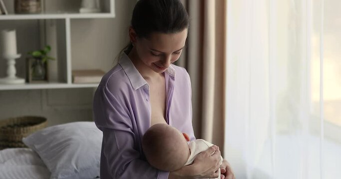 Young 25s beautiful mother breastfeeds newborn baby seated on bed at home. Tender moment of feed, healthy nutrition for health and growth of infant child, lactation, care, babyhood development concept