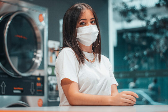 Adolescente triste joven de pelo negro con mascarilla esperando junto a las maquinas de lavado industrial en la lavanderia del comercio para recoger su ropa lavada desinfectada y limpia