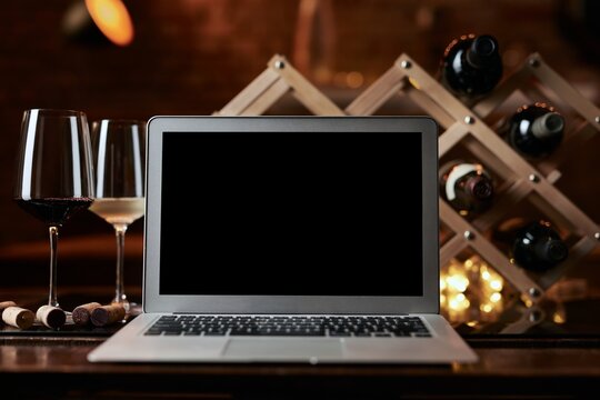 Empty blank screen laptop computer on table with wine bottle wine rack holder in the background. Buying wines online, home delivery concepts.