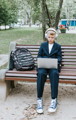 cute schoolboy with backpack in a school uniform with headphones and laptop is sitting on bench in the school yard, concept back to school and start of lessons