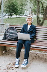 Fototapeta premium cute schoolboy with backpack in a school uniform with headphones and laptop is sitting on bench in the school yard, concept back to school and start of lessons