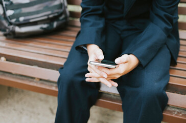 cute schoolboy with backpack in a school uniform with wireless headphones and using mobile phone is sitting on bench in the school yard, concept back to school and start of lessons