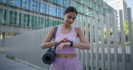Sportive woman manages clock on smartwatch tracks time checks health indicators on electronic wristwatch installs modern application to see fitness results dressed in activewear stands in downtown - Powered by Adobe