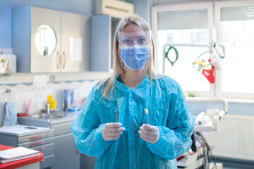 Female dentist in a protective suit with a protective shield and mask holding dental tools in dental office. - obrazy, fototapety, plakaty