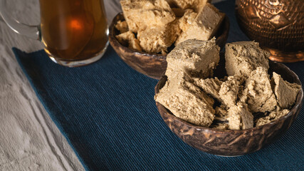 Sunflower halva is in a ceramic bowl on a blue napkin, there is a copper vase and a cup of tea at the back. Traditional oriental sweetness for tea drinking. Close-up, copying the space..