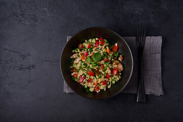Traditional salad tabbouleh made of bulgur or couscous, poultry meat, parsley, mint in dark bowl with napkin and fork on black background