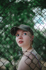 a girl with short hair in a baseball cap, looking through a mesh fence, through a mesh, bright blue eyes