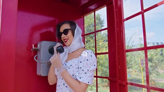 beautiful young woman happily speaks on the phone in an english style red telephone booth. girl dressed in a white dress and glasses. pinup.