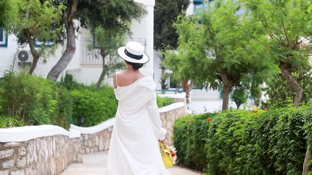 Cinematic slow motion Young woman in white dress walks holding a hat walking through the streets of greece drone flies overhead