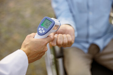 Close up shot of medical infrared thermometer in a hand of doctor measuring the temperature of male handicapped patient in wheelchair during Covid19 outbreak