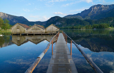 a pier leading to the wooden boat houses on serene lake Kochel (Kochelsee) with the scenic Bavarian Alps in the background reflected in the water (Bavaria, Germany)