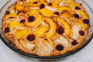 Delicious, fresh, baked pie of apples, piers, raspberries, apricot close-up in a round glass form stands on a white table.